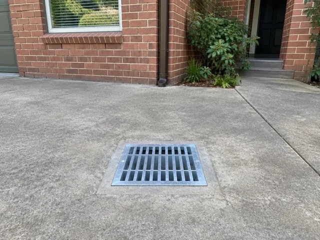 Concrete driveway with a square metal drain grate beside a brick house in Wahroonga